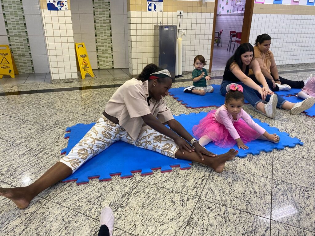 Crianças e adultos estão sentados em tapetes azuis, esticando-se juntos em um piso de ladrilho. O adulto na frente está guiando uma jovem garota em com roupa de Ballet.