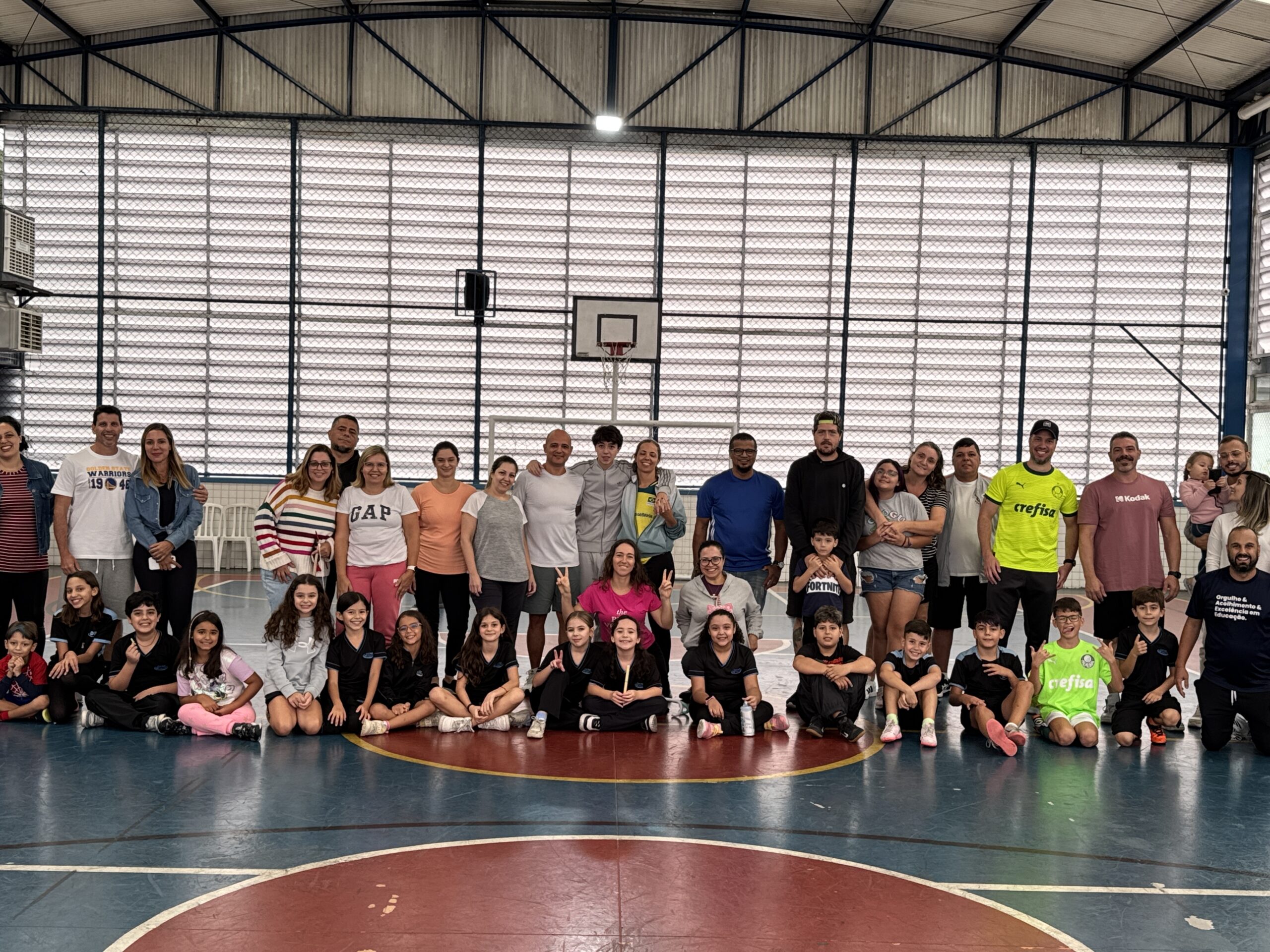 Grupo grande de adultos e crianças posando para foto em uma quadra esportiva coberta. As crianças estão sentadas na frente, muitas com uniformes esportivos, enquanto os adultos estão de pé ao fundo, sorrindo. Ao fundo, há uma tabela de basquete e paredes de grade com iluminação natural entrando. A atmosfera é de confraternização e alegria.