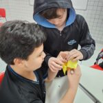 Estudantes do 5th Grade do Colégio Dom Bosco, uniformizados, participam de uma atividade em sala de aula. Eles estão sentados em grupos, interagindo com papéis coloridos dobrados em formato de origami da sorte. As expressões dos alunos revelam concentração e entusiasmo enquanto exploram perguntas em inglês com as Wh-words. O ambiente é acolhedor e descontraído, reforçando o caráter lúdico e colaborativo da atividade.