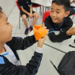 Estudantes do 5th Grade do Colégio Dom Bosco, uniformizados, participam de uma atividade em sala de aula. Eles estão sentados em grupos, interagindo com papéis coloridos dobrados em formato de origami da sorte. As expressões dos alunos revelam concentração e entusiasmo enquanto exploram perguntas em inglês com as Wh-words. O ambiente é acolhedor e descontraído, reforçando o caráter lúdico e colaborativo da atividade.