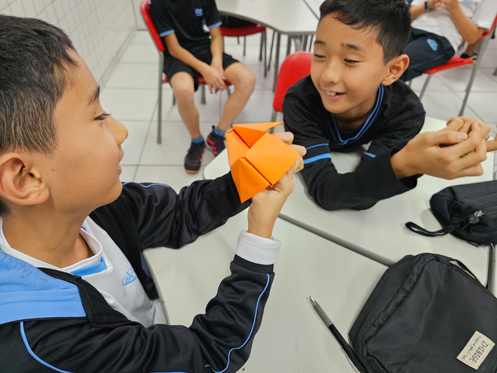 Estudantes do 5th Grade do Colégio Dom Bosco, uniformizados, participam de uma atividade em sala de aula. Eles estão sentados em grupos, interagindo com papéis coloridos dobrados em formato de origami da sorte. As expressões dos alunos revelam concentração e entusiasmo enquanto exploram perguntas em inglês com as Wh-words. O ambiente é acolhedor e descontraído, reforçando o caráter lúdico e colaborativo da atividade.
