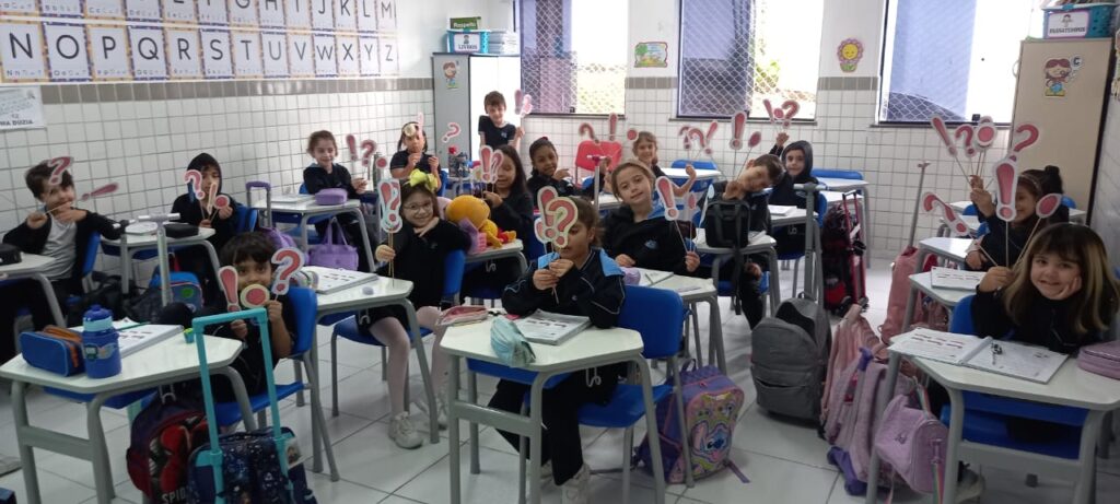 Alunos do 2º ano segurando cartazes com sinais de pontuação em sala de aula, participando de atividade lúdica sobre o uso do ponto final, exclamação e interrogação.