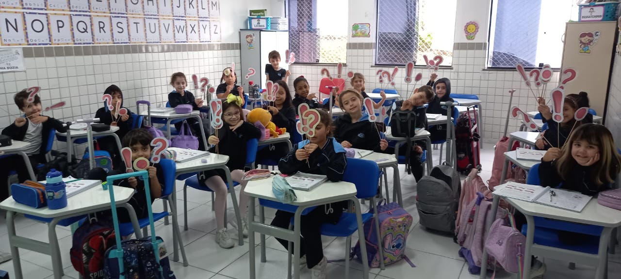 Alunos do 2º ano segurando cartazes com sinais de pontuação em sala de aula, participando de atividade lúdica sobre o uso do ponto final, exclamação e interrogação.