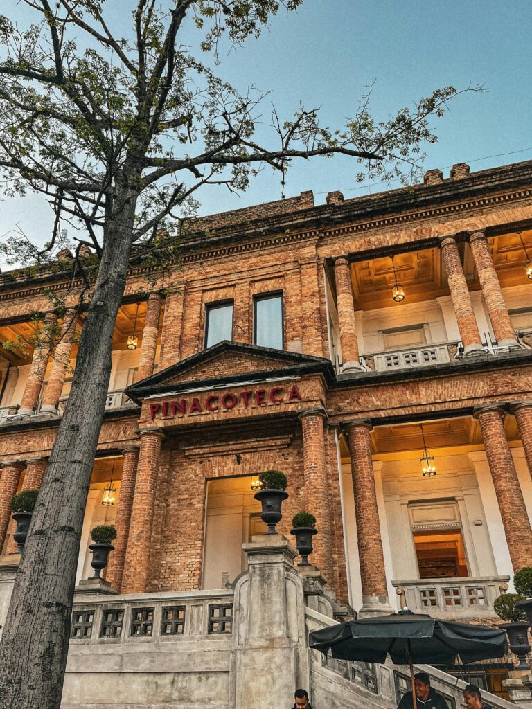 Fachada principal da Pinacoteca de São Paulo ao entardecer, com sua arquitetura de tijolos aparentes, colunas imponentes e escadaria frontal.