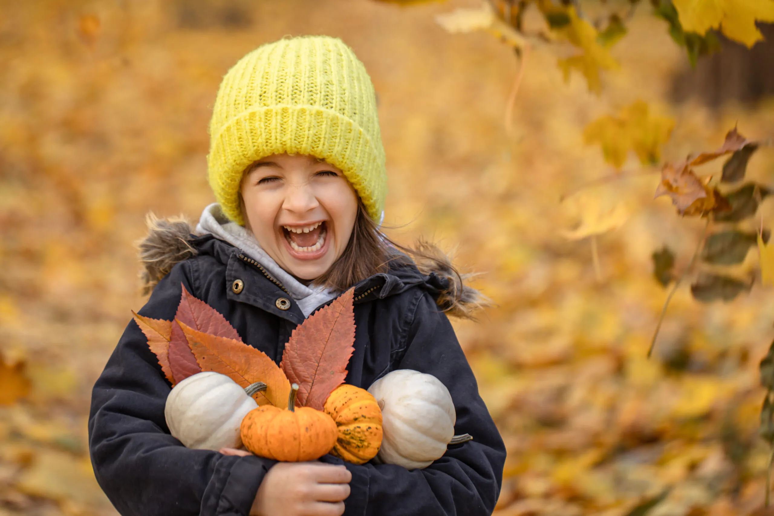 Uma criança alegre usando um gorro de tricô amarelo e um casaco escuro, segurando pequenas abóboras e folhas coloridas de outono. O fundo é preenchido com folhas caídas e árvores em foco suave, criando uma atmosfera quente de outono.