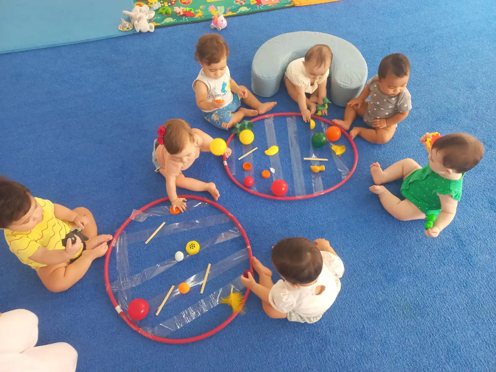 Bebês do Berçário do Colégio Dom Bosco sentados em círculo sobre tapete azul, explorando brinquedos coloridos colados em bambolês com fitas adesivas, em atividade sensorial de estímulo à coordenação motora e curiosidade.