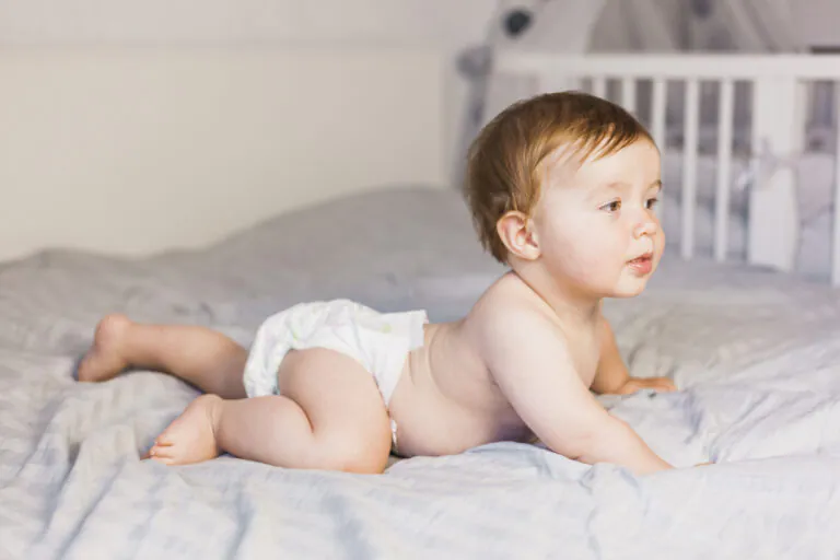 Bebê de fralda deitado de bruços na cama, em fase inicial do desfralde, com berço ao fundo em ambiente acolhedor.