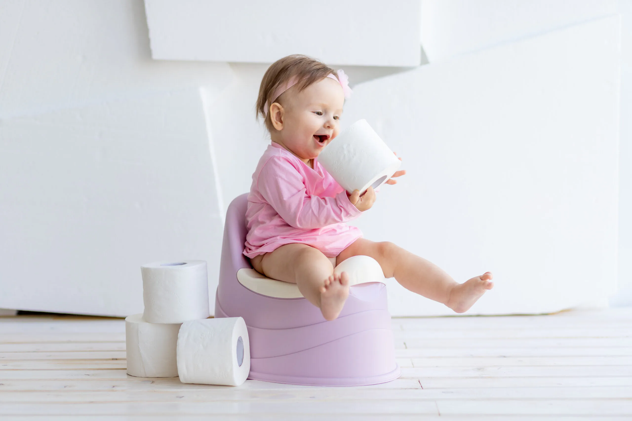 Bebê sorridente sentada em um penico lilás, vestindo roupa rosa e segurando um rolo de papel higiênico, com outros rolos empilhados ao lado, em um ambiente claro e minimalista.