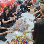 Uma visão de cima mostra um grupo de estudantes sentados no chão de uma sala de aula clara e bem iluminada. Eles estão cercados por uma grande quantidade de embalagens de doces coloridas, espalhadas pelo chão. Os estudantes parecem estar envolvidos em alguma atividade relacionada aos doces.