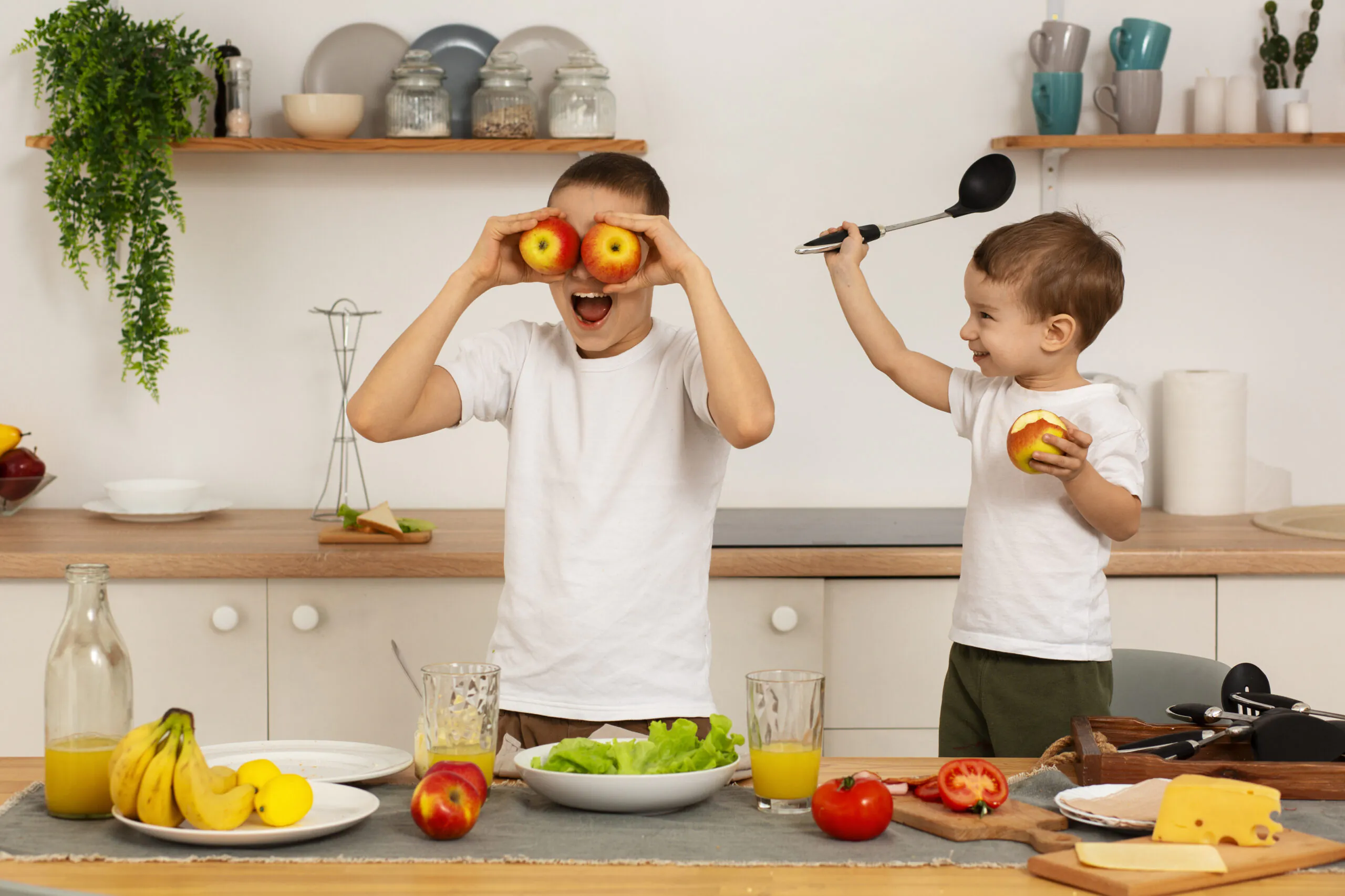 Duas crianças se divertem na cozinha enquanto preparam uma refeição saudável. Uma segura duas maçãs nos olhos como se fossem óculos, e a outra sorri segurando uma maçã e uma concha de cozinha. Em cima da mesa há frutas, salada, suco, queijo e utensílios de cozinha.