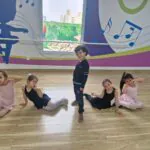 Crianças sorrindo em pose na aula de de Ballet Infantil, vestindo figurinos em tons de rosa e preto, participando de atividade lúdica de dança em ambiente escolar decorado com notas musicais coloridas na parede.