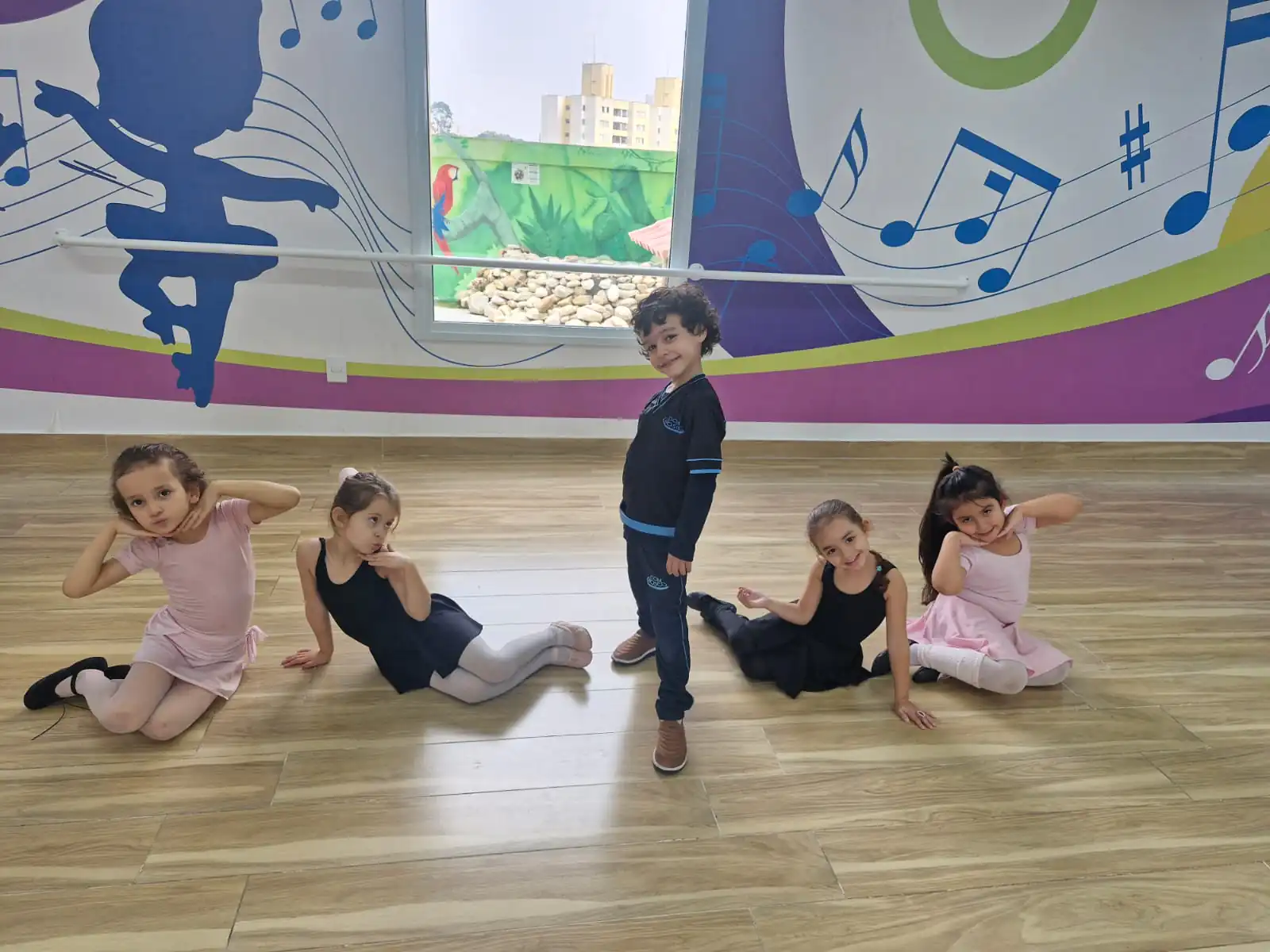Crianças sorrindo em pose na aula de de Ballet Infantil, vestindo figurinos em tons de rosa e preto, participando de atividade lúdica de dança em ambiente escolar decorado com notas musicais coloridas na parede.