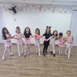 Crianças sorrindo e de mãos dadas em sala de aula de Ballet Infantil, vestindo figurinos em tons de rosa e preto, participando de atividade lúdica de dança em ambiente escolar decorado com notas musicais coloridas na parede.