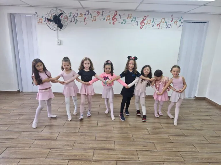 Crianças sorrindo e de mãos dadas em sala de aula de Ballet Infantil, vestindo figurinos em tons de rosa e preto, participando de atividade lúdica de dança em ambiente escolar decorado com notas musicais coloridas na parede.
