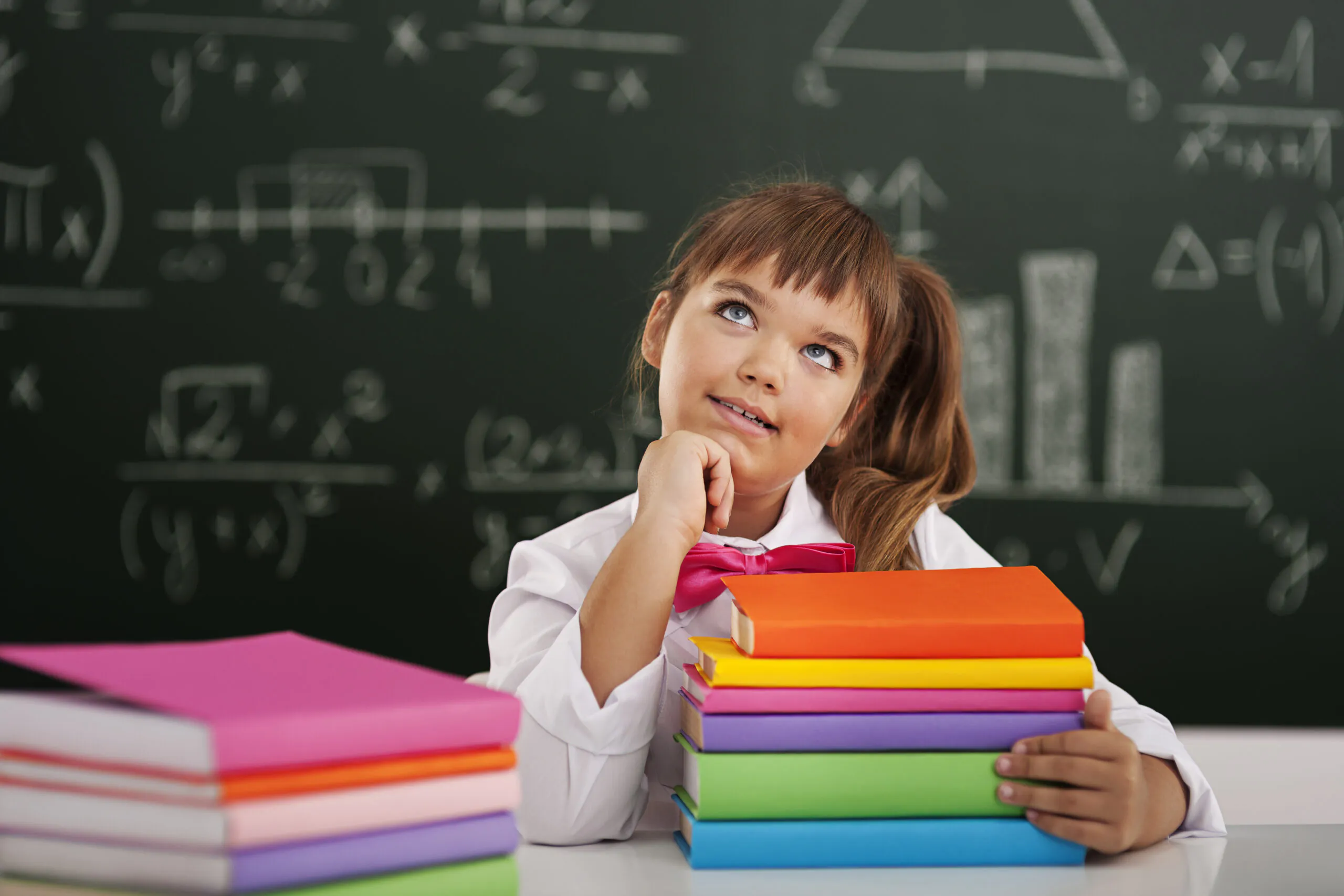 Menina sorridente com uniforme escolar e laço rosa, segurando uma pilha de livros coloridos em sala de aula com quadro-negro ao fundo cheio de fórmulas matemáticas. Conceito de volta às aulas e transição para o Ensino Fundamental.