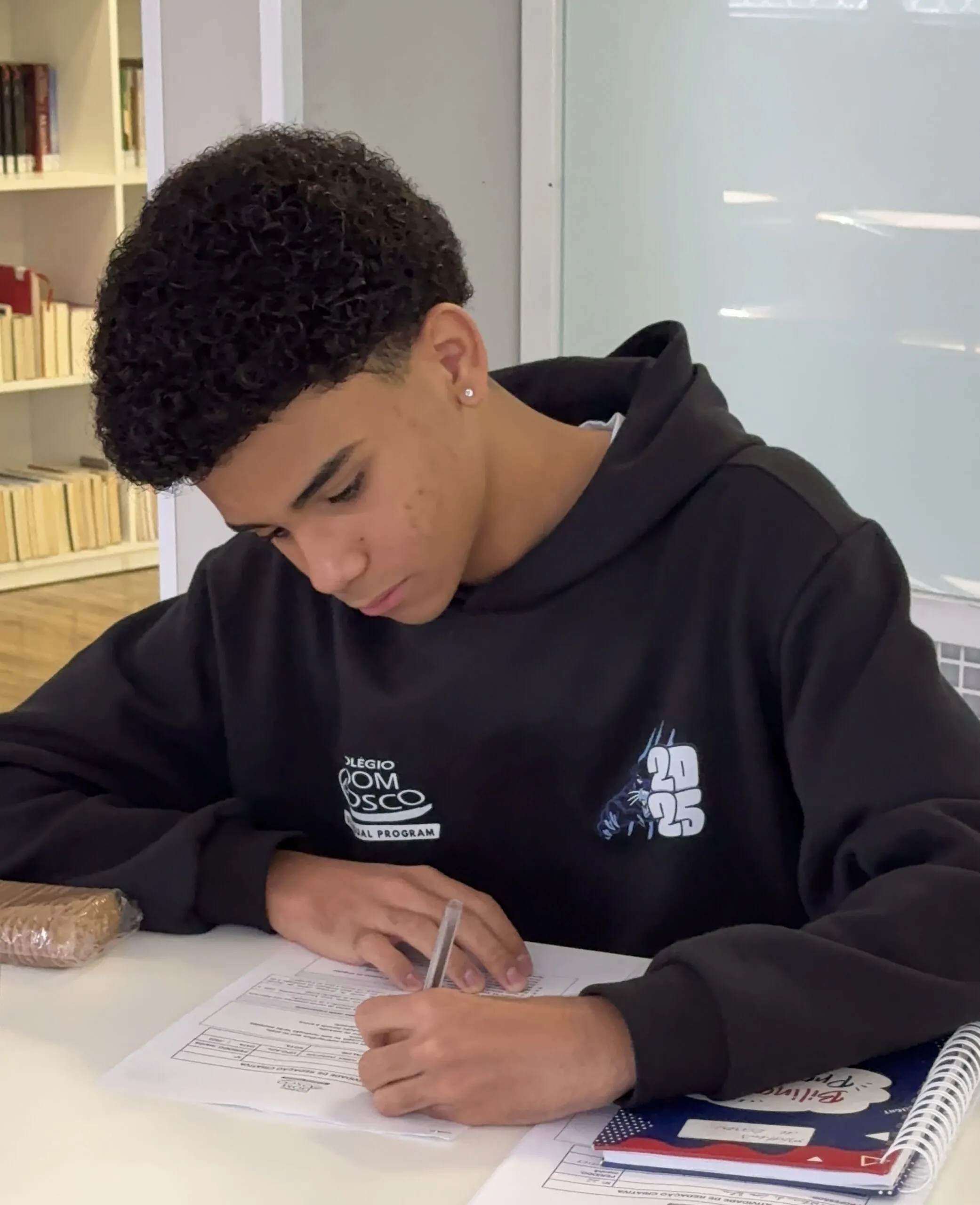 Estudante do Colégio Dom Bosco, vestindo moletom preto com o logo da escola e o número 2025, está concentrado preenchendo uma folha de atividades em uma sala de aula. Sobre a mesa, há um caderno, uma apostila e um pacote de bolachas.